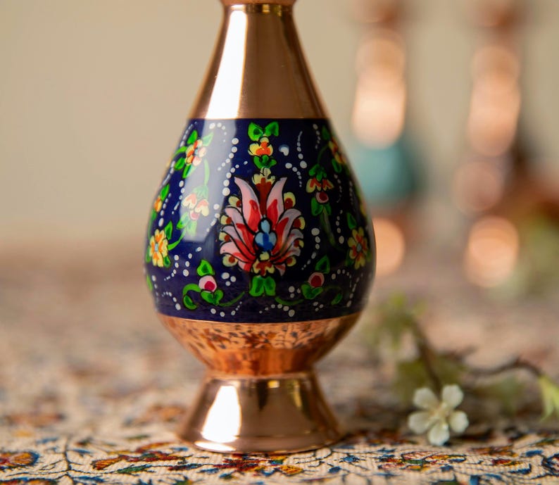 May include: A copper vase with a navy blue center section adorned with a floral design in red, pink, yellow, and green. The vase has a flared base and a narrow neck, showcasing a blend of metallic and painted elements.