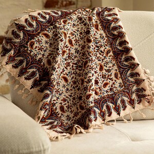 May include: A beige throw blanket with a detailed paisley pattern in burgundy, navy blue, and cream. The blanket has a fringed edge and is draped over a light-colored sofa, showcasing its intricate design.