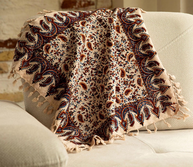 May include: A beige throw blanket with a detailed paisley pattern in burgundy, navy blue, and cream. The blanket has a fringed edge and is draped over a light-colored sofa, showcasing its intricate design.