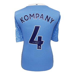 以下が含まれることがあります： 背中に「KOMPANY」の名前と番号「4」が入った水色のサッカーユニフォーム。ユニフォームには白い襟があり、襟の後ろには「CITY」の文字がプリントされています。袖には白と水色の模様があります。