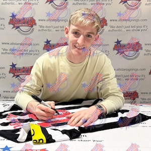 May include: A person signing a black and white sports jersey with a black marker. The jersey has red accents. The background features a repeating logo with the text "Allstar Signings".
