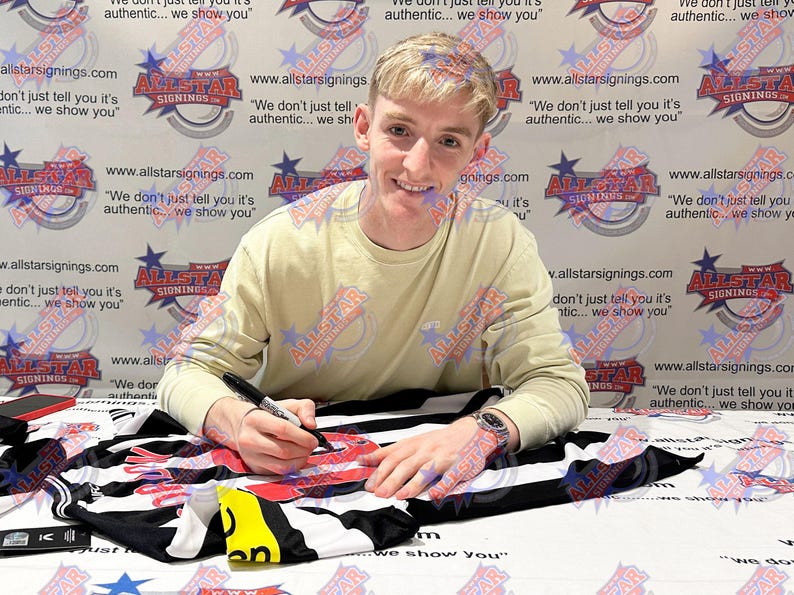 May include: A person signing a black and white sports jersey with a black marker. The jersey has red accents. The background features a repeating logo with the text "Allstar Signings".