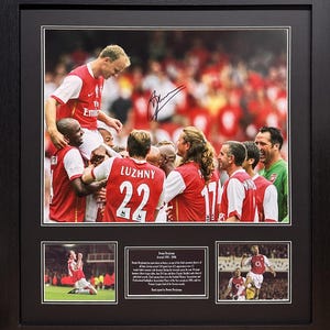 May include: A framed photo collage of a soccer player celebrating with his team. The main photo shows the player being lifted up by his teammates. The other photos show the player scoring a goal and celebrating. The photo is signed by the player, Dennis Bergkamp.