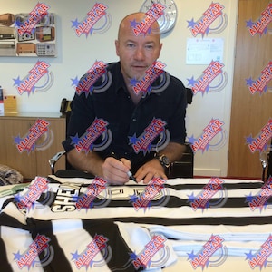 May include: A man in a black shirt is signing a black and white striped jersey with the word "SHE" printed on it.