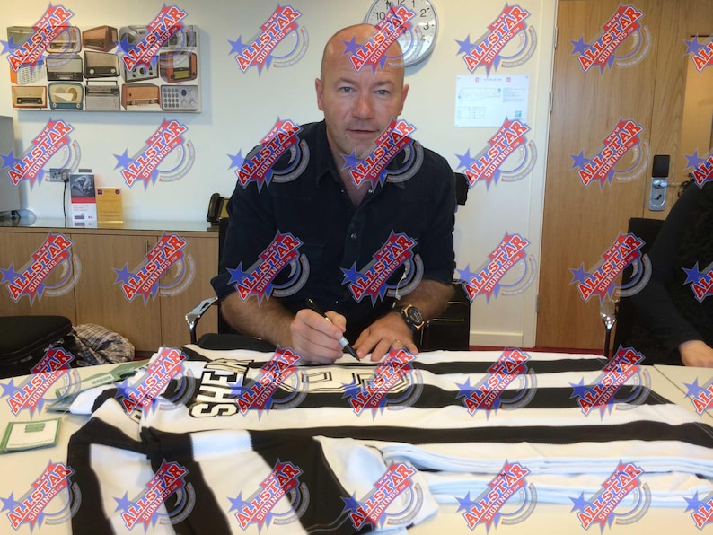 May include: A man in a black shirt is signing a black and white striped jersey with the word "SHE" printed on it.