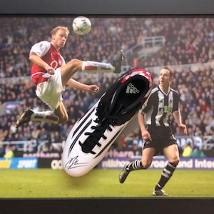 May include: A white and black soccer cleat with a red stripe and the Adidas logo. The cleat is signed by a soccer player. The cleat is displayed in a framed photo of a soccer player kicking a ball.