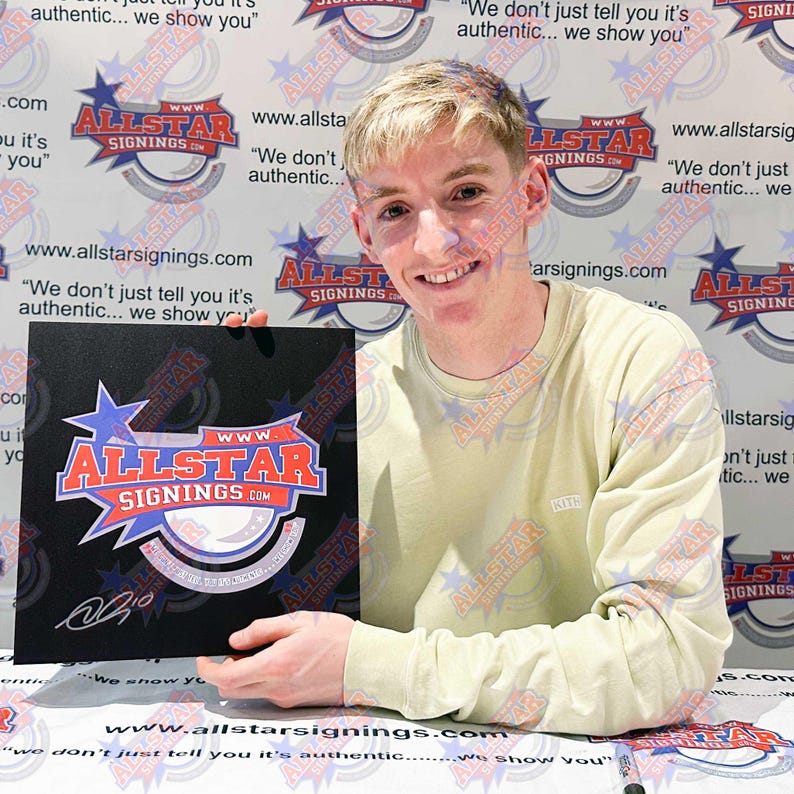 May include: A person holding a black square with the "ALLSTAR SIGNINGS" logo in red, white, and blue. The background features the same logo and text. The person is wearing a light green long-sleeve shirt.