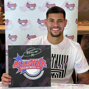 May include: A person holding a black square with the Allstar Signings logo and signature. The person is wearing a white t-shirt with a black and white graphic design. The background features the Allstar Signings logo.