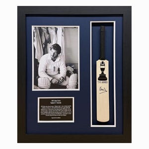 Puede incluir: Una foto en blanco y negro enmarcada de un jugador de cricket con un uniforme blanco, sosteniendo un bate de cricket. La foto está al lado de un bate de cricket en miniatura enmarcado con el texto "The Ashes" y una firma. El marco es negro con un passe-partout azul.
