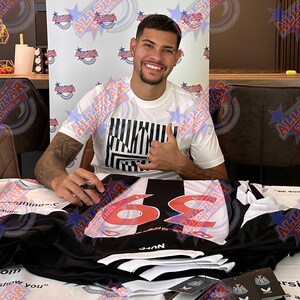 May include: A person smiling and giving a thumbs-up, seated behind a table. A black and white soccer jersey with the number 69 in red is on the table. The person is wearing a white t-shirt with a black and white graphic.