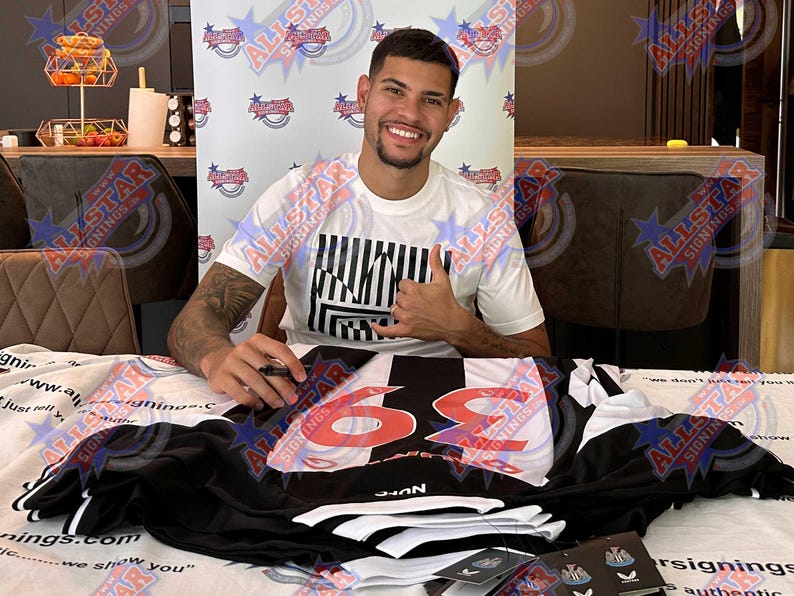 May include: A person smiling and giving a thumbs-up, seated behind a table. A black and white soccer jersey with the number 69 in red is on the table. The person is wearing a white t-shirt with a black and white graphic.