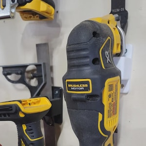 Op de afbeelding: Een close-up van een zwart en geel draadloos oscillerend gereedschap met een witte wandbevestiging. Het gereedschap heeft een zwarte handgreep en een gele trekker. Het gereedschap is gelabeld met "BRUSHLESS MOTOR" en "XR".