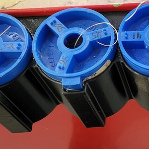 Può includere: Tre bobine di plastica blu con filo, ciascuna con un'etichetta diversa: "Safety", "Lock Wire" e "Safety". Le bobine sono montate su un supporto in plastica nera.