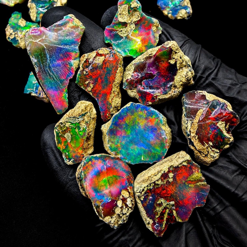 May include: A collection of raw, unpolished opal stones featuring vibrant flashes of iridescent color including red, green, blue, and yellow. Each stone displays a unique, irregular shape with rough, textured edges and a polished, reflective surface.