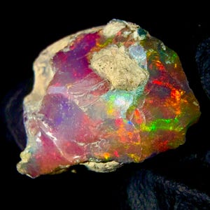 May include: A raw, unpolished opal gemstone displaying a vibrant play of color with flashes of neon red, bright green, and deep purple against a translucent, milky base. The stone features an irregular, jagged shape with a rough, beige matrix.