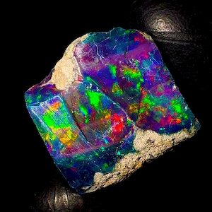 May include: A vibrant, raw opal gemstone with a striking play of color. The stone displays a spectrum of hues, including blues, greens, purples, and reds, set against a dark background. The opal's surface has a rough, natural texture.