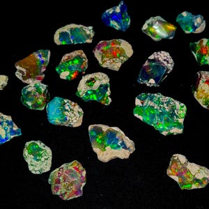 May include: A collection of rough opal gemstones, each displaying vibrant flashes of color including blues, greens, and reds. The opals are set against a black background, highlighting their natural, unpolished state. The stones vary in size and shape, showcasing their unique characteristics.