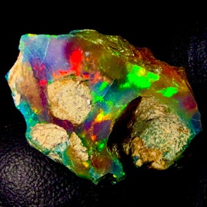 May include: A raw, unpolished opal gemstone featuring a vibrant play of color with flashes of neon green, bright red, and deep blue. The stone has an irregular, jagged shape with embedded patches of light tan, textured, non-iridescent rock matrix.