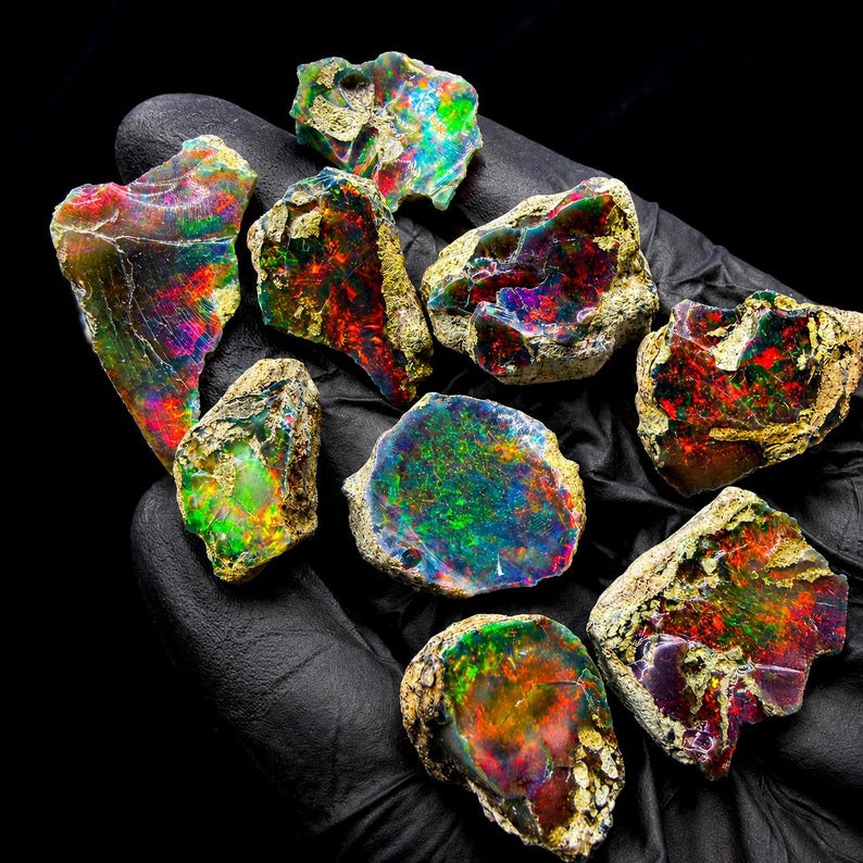 May include: A collection of nine raw, unpolished opal stones featuring vibrant flashes of red, green, blue, and purple color play. Each stone displays a unique, irregular shape with a rough, textured exterior and a polished, iridescent face.