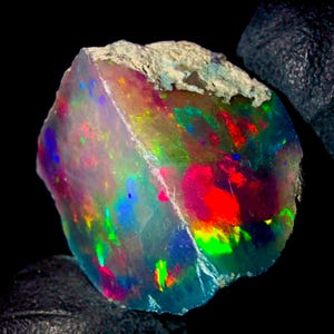 May include: A raw, uncut opal gemstone featuring a vibrant play of colour with flashes of neon red, electric blue, and bright green against a translucent, milky base. The stone has a rough, textured, light-coloured mineral crust at the top edge.