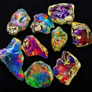 May include: A collection of nine raw, unpolished opal stones featuring vibrant flashes of red, blue, green, and purple iridescence. Each stone displays a unique, irregular shape with rough, textured edges and a mix of colorful, glossy surfaces.