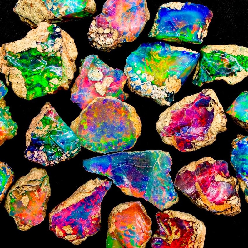 May include: A collection of raw, unpolished opal stones featuring vibrant flashes of neon green, electric blue, fiery red, and bright orange. Each irregular rock fragment displays a unique play-of-color against a natural, sandy-colored matrix.
