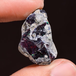 Könnte beinhalten: Ein rauer, dunkelgrauer und schwarzer Opal mit irisierenden Schimmern in Grün und Rot. Der Opal ist ein Naturstein und hat eine raue, unpolierte Oberfläche.
