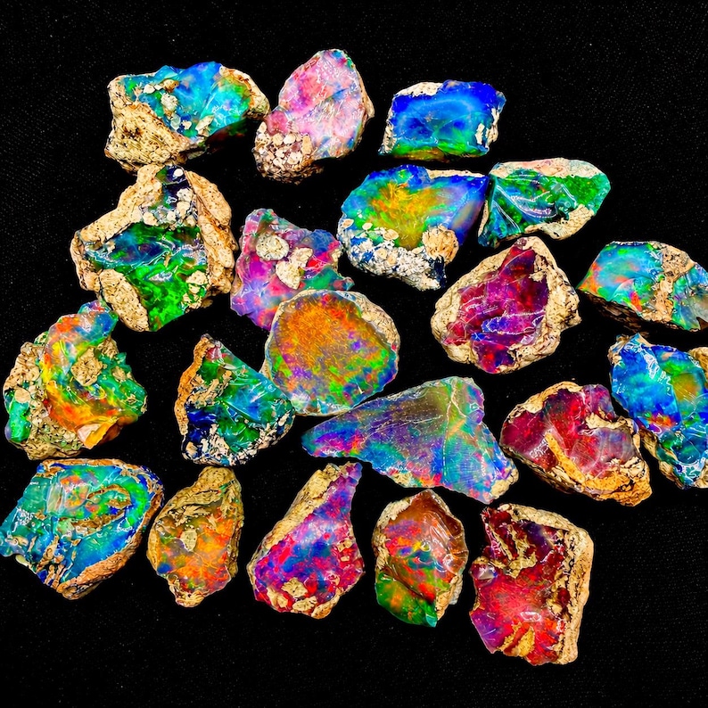 May include: A collection of raw, unpolished opal stones featuring vibrant flashes of iridescent color including neon green, electric blue, fiery red, and bright orange, with rough, textured, tan-colored rock matrix visible on the edges of each piece.