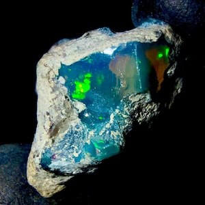 Könnte beinhalten: Ein roher, ungeschliffener Opal-Edelstein mit einem lebendigen Farbspiel. Der Opal zeigt grüne und blaue Blitze in einer Matrix aus braunem und grauem Gestein. Der Edelstein ist vor einem schwarzen Hintergrund platziert, was seine natürliche Schönheit hervorhebt.