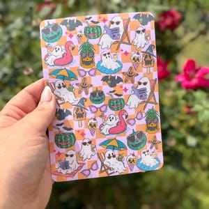 May include: A rectangular sticker sheet featuring a repeating checkered pattern in light pink and orange. The design includes various illustrations of ghosts wearing sunglasses, pineapples, bats, flamingos, and drinks with the text Spooky Summer.