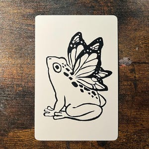 May include: A black and white illustration of a frog with butterfly wings. The frog is sitting on a white rectangular card. The butterfly wings have a detailed pattern. The card is on a wooden surface.