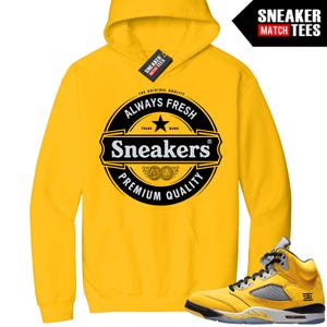 May include: A yellow hoodie with a graphic that reads "Always Fresh Sneakers Premium Quality." A pair of yellow and gray sneakers is displayed next to the hoodie. The hoodie has a tag that says "Sneaker Match Tees."