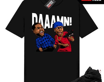 May include: Black t-shirt featuring a cartoon graphic of two men with the word "DAAAMN!" above them. One man wears a blue plaid shirt, the other a red shirt and blue cap. A black and red sneaker is shown below the shirt.