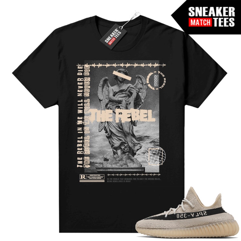K&ouml;nnte beinhalten: Schwarzes T-Shirt mit einer Grafik einer Statue und den Worten "THE REBEL" und "THE REBEL IN WE WILL NEVER DIE". Ein Paar beige-schwarze Turnschuhe befindet sich im Vordergrund. Das Shirt hat ein Etikett mit der Aufschrift "SNEAKER MATCH TEES".