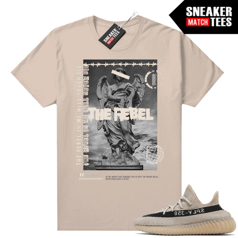 May include: A beige t-shirt with a graphic of a statue and the words "THE REBEL". A pair of beige and black trainers are in the bottom right corner. The shirt has a tag that says "SNEAKER MATCH TEES".