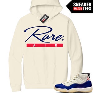 Puede incluir: Sudadera con capucha color crema con el texto "Rare Air" en escritura azul marino y letras rojas. Un par de zapatillas deportivas azules, blancas y negras se muestran junto a la sudadera con capucha. La sudadera tiene una capucha con cordón y una etiqueta que dice "Sneaker Match Tees".