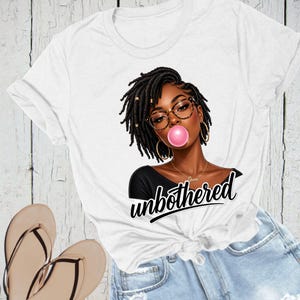 Puede incluir: Camiseta blanca con un gr&aacute;fico de una mujer negra con rastas, gafas y una burbuja de chicle rosa. La palabra "unbothered" est&aacute; escrita en cursiva debajo del gr&aacute;fico. La camiseta est&aacute; anudada a la cintura.