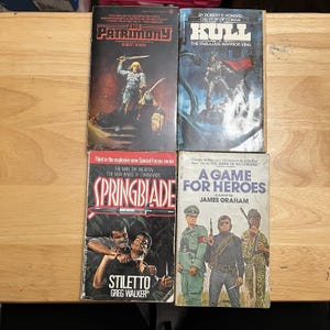 May include: Four paperback books arranged in a grid. Top left: The Patrimony by Robert Adams. Top right: Kull The Fabulous Warrior King by Robert E. Howard. Bottom left: Springblade Stiletto by Greg Walker. Bottom right: A Game for Heroes by James Graham.