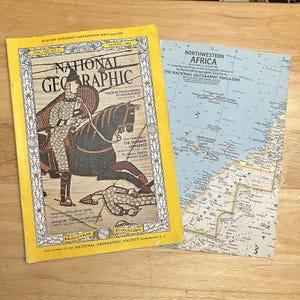 Puede incluir: Dos revistas National Geographic antiguas. El número de agosto de 1966 presenta una ilustración de portada de un caballero a caballo. La otra es un suplemento de mapa del noroeste de África. Ambas están sobre una superficie de madera clara.