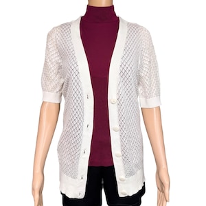 May include: A white, short-sleeved cardigan with a button-up front. The cardigan has a delicate, open-weave design. The model is wearing a burgundy turtleneck and black shorts. The cardigan is a versatile layering piece.