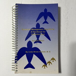 Könnte beinhalten: Ein spiralgebundenes Handbuch mit dem Titel "Symptom Management Algorithms: A Handbook for Palliative Care, Second Edition." Das Cover ist blau mit stilisierten blauen Vogelgrafiken und goldfarbenen Buchstaben. Die Autorin ist Linda Wrede-Seaman, M.D.