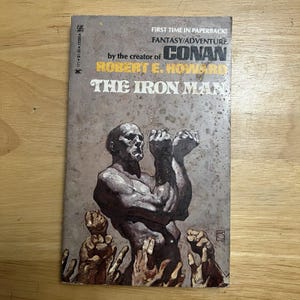 Könnte beinhalten: Ein Vintage-Taschenbuch mit dem Titel "The Iron Man" von Robert E. Howard. Das Cover zeigt eine muskulöse Figur mit erhobenen Fäusten und mehreren Händen, die sich erheben. Das Buch stammt vom Zebra-Verlag.
