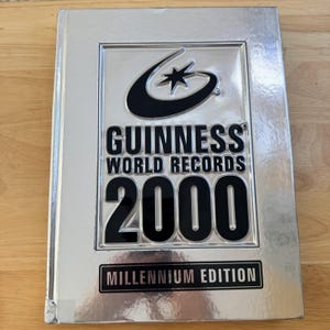 Peut inclure: Un livre Guinness World Records 2000 de couleur argentée. La couverture présente le logo Guinness, le texte "GUINNESS WORLD RECORDS 2000" et "MILLENNIUM EDITION". Le livre est sur une surface en bois.