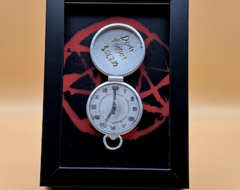 May include: A framed artwork featuring a silver pocket watch design. The watch is open, revealing the time. The background has a red pentagram design on a black surface. The top of the watch has the words "Don't Forget 3.OCT.10".