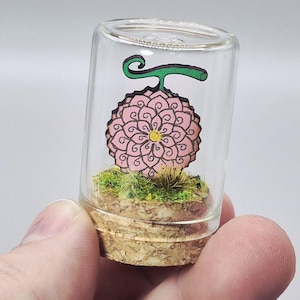 May include: A miniature glass terrarium featuring a pink, flower-like design with black outlines and a green stem. The terrarium is filled with green moss and sits atop a cork base. The clear glass allows full view of the detailed artwork.
