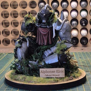 May include: A detailed figurine of Alphonse Elric from Fullmetal Alchemist, featuring a metallic gray armored body with red accents and green moss detailing. The figurine is mounted on a circular base with a nameplate that reads "Alphonse Elric Fullmetal Alchemist."