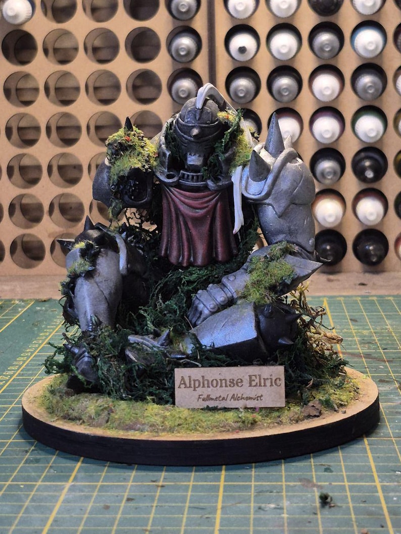 May include: A detailed figurine of Alphonse Elric from Fullmetal Alchemist, featuring a metallic gray armored body with red accents and green moss detailing. The figurine is mounted on a circular base with a nameplate that reads "Alphonse Elric Fullmetal Alchemist."