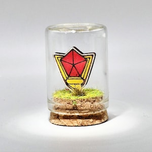 May include: A small glass bottle with a cork base, containing a miniature landscape. Inside, a wooden emblem with red and yellow accents sits atop a patch of green moss. The emblem has a geometric design. The bottle is clear, allowing full view of the contents.