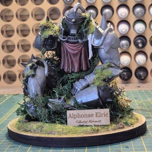 May include: A detailed figurine of Alphonse Elric from Fullmetal Alchemist, crafted with metallic armor and a red cloth. The figure is surrounded by green moss and sits on a wooden base with a nameplate.