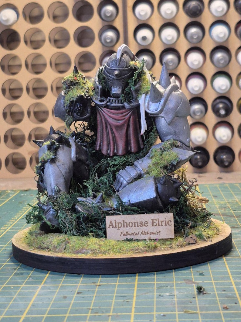 May include: A detailed figurine of Alphonse Elric from Fullmetal Alchemist, crafted with metallic armor and a red cloth. The figure is surrounded by green moss and sits on a wooden base with a nameplate.